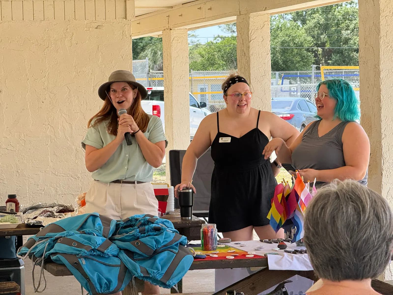 Three speakers laughing together at the front of a pavilion picnic, with Pride flags on the table.