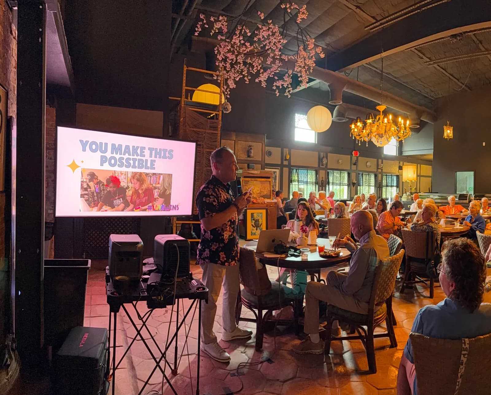 A speaker at the front of a restaurant full of supporters, beside a screen that reads You Make This Possible.