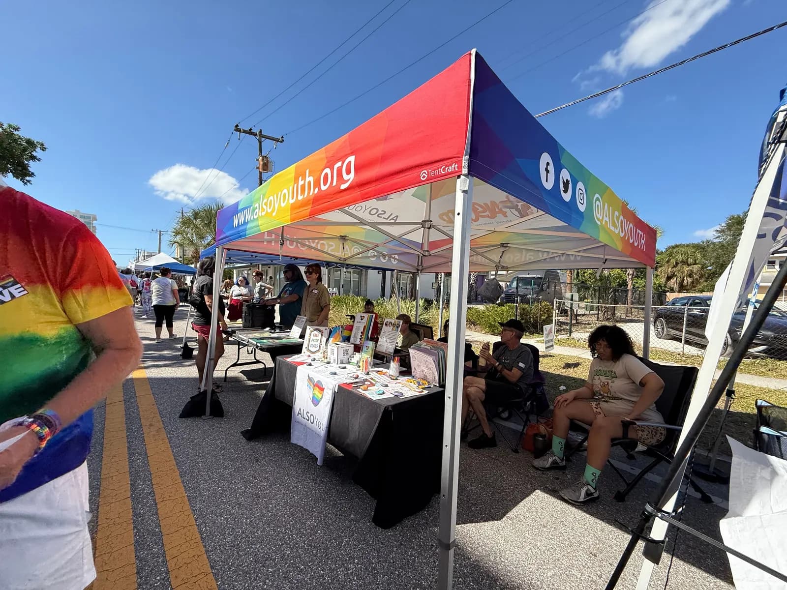 The ALSO Youth rainbow tent on a closed street under a bright blue sky, with the booth set up and people passing by.